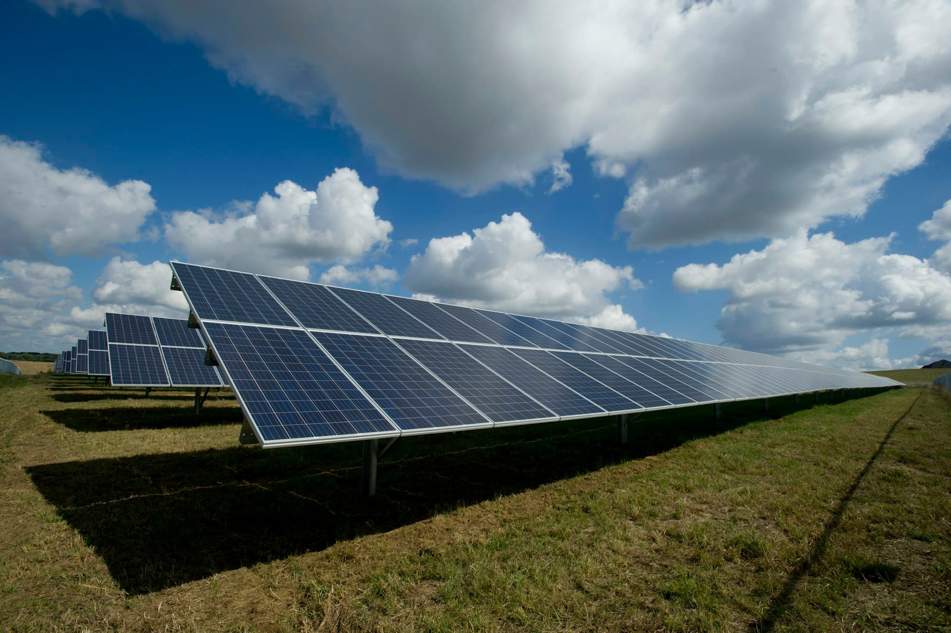 Wide angle shot of a massive solar farm field with a technician in safety gear inspecting a panel under a clear blue sky